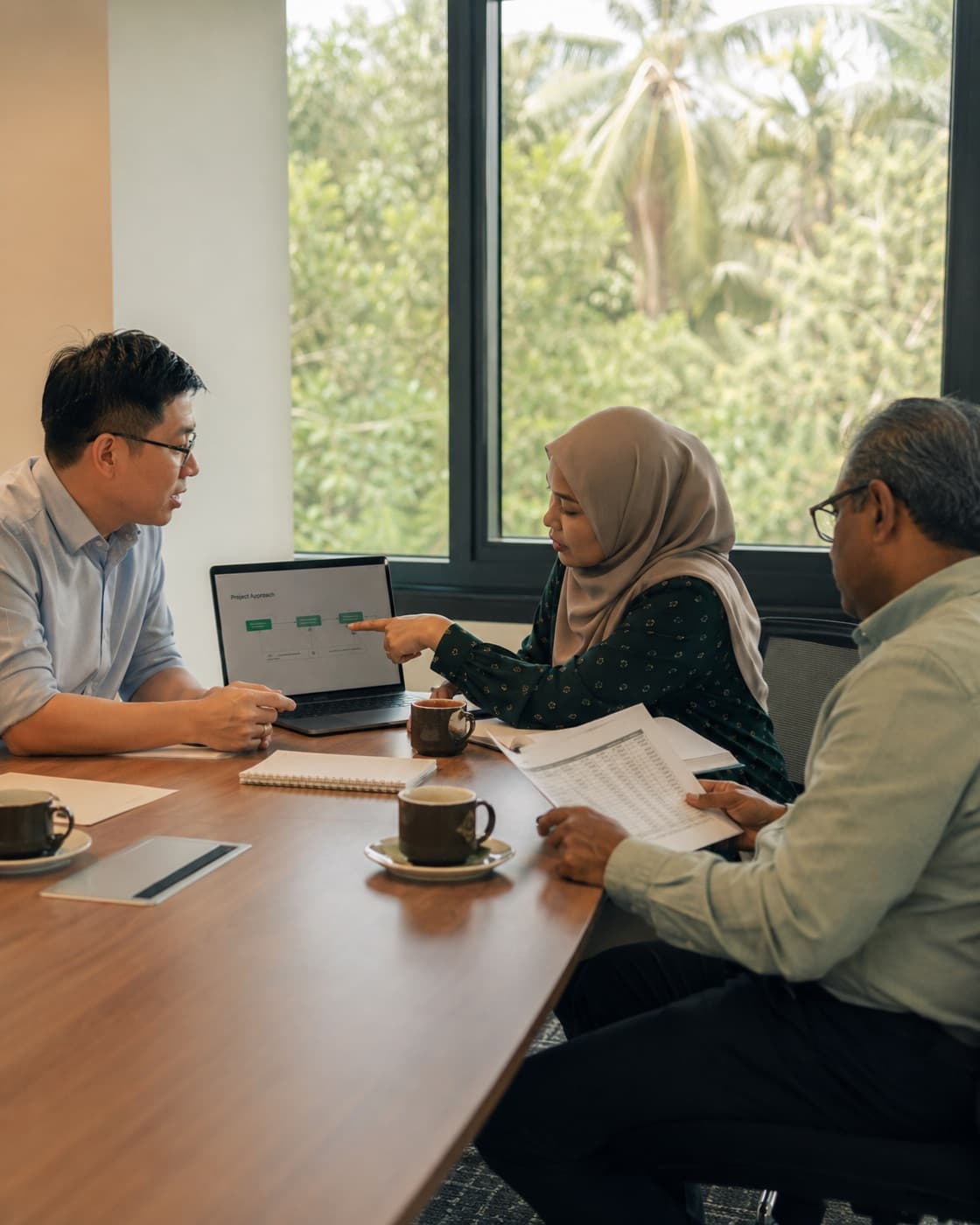 Three people around a meeting table, one pointing at a project approach diagram on a laptop while another holds a printed report.