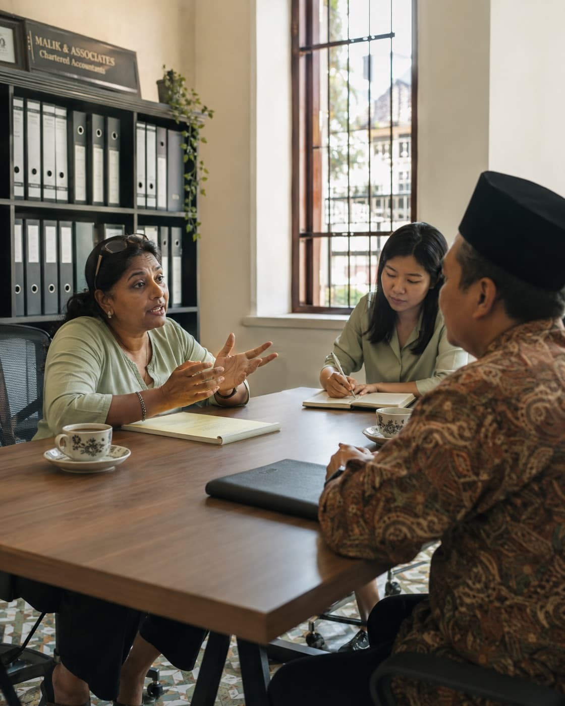 An accountant explaining a point across a meeting table to a client and a colleague, box files on the shelf behind.